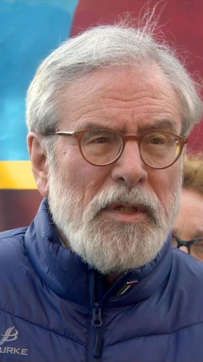 Gerry Adams is speaking into the microphones and is wearing a blue gillet and a light blue shirt. He is standing in front of a mural of Bobby Sands.