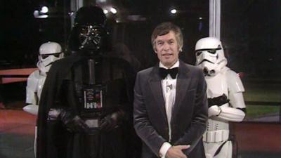 David Prowse as Darth Vader standing next to Jackie Collins with stormtroopers standing behind them.