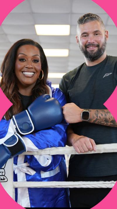 Alison Hammond and Tony Bellew stand on the edge of a boxing ring. Alison is wearing a blue boxing glove and fist pumping Tony.