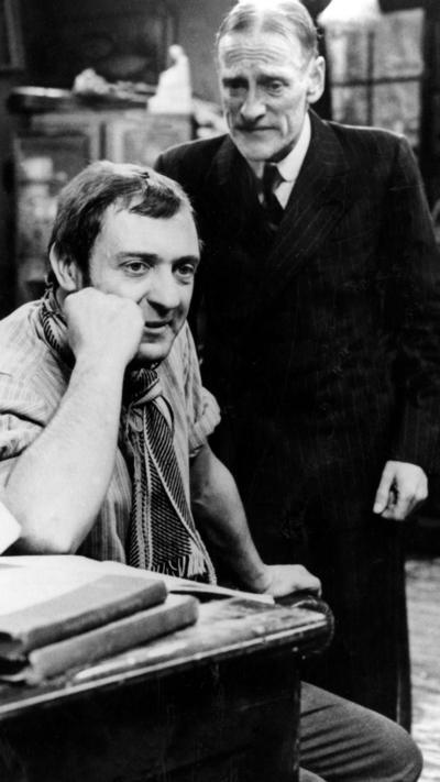 A black and white still from TV sit com Steptoe and Son. A young man sits at a desk resting his head on his hand. He is wearing a striped top and neck scarf. Behind him an older man with white hair stands looking towards him. He is wearing a dark suit, white short and tie.
