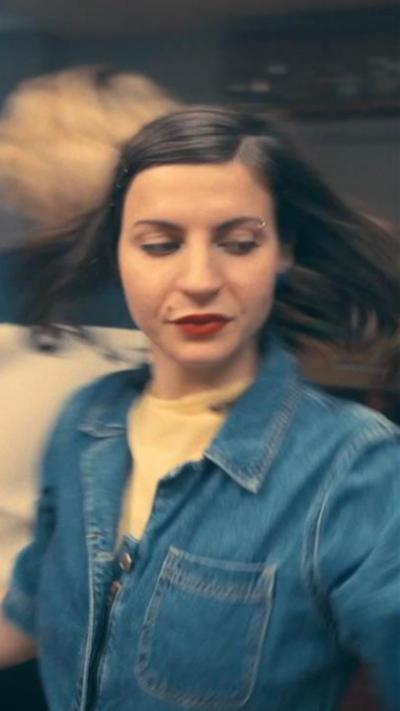 A woman in a short-sleeved, collared denim shirt with a yellow T-shirt underneath is dancing. Her hair is in motion as she spins, flowing out on each side of her head. She is looking down and to to her left.