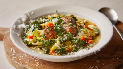 A white bowl with veggie meatball orzo soup in. A silver spoon sits next to the bowl. 