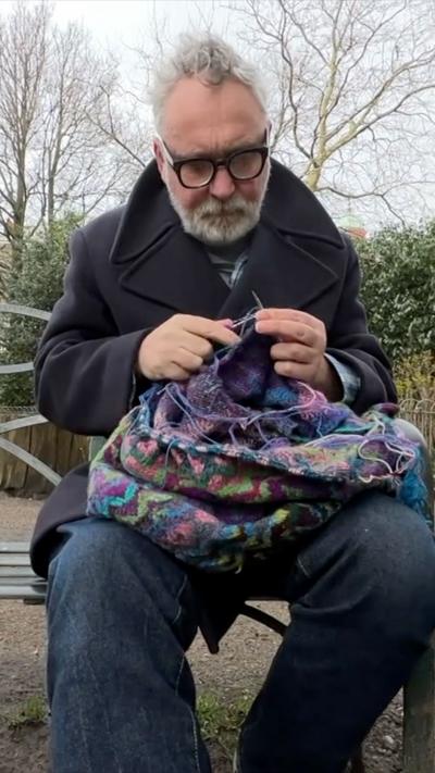 Elderly man with white hair and beard knitting on a bench outdoors