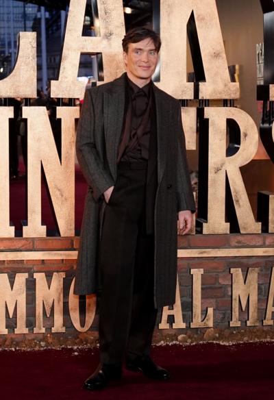 Man dressed in a grey long coat with burgundy shirt and scarf and black trousers. Stands in front of a huge sign that says Peaky Blinders. His hand is in his pocket as he stares into camera.