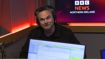 A man with headphones on in a radio studio. In the foreground, in front of him, we can see the front of a computer screen used by the radio programme presenter. In the background is a screen showing the BBC News Northern Ireland logo