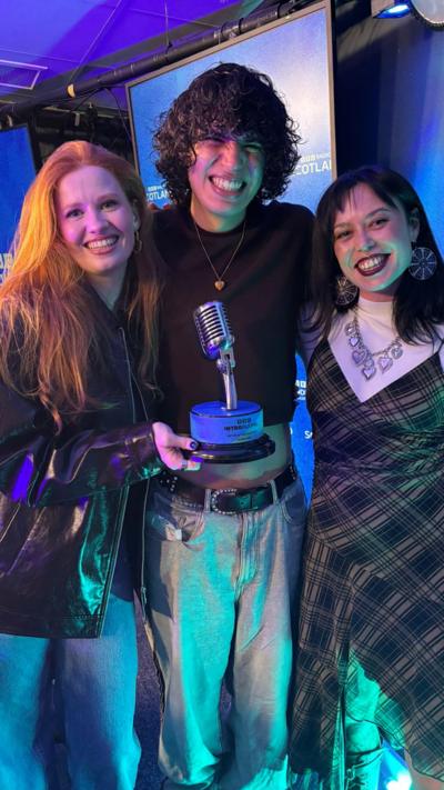 A woman with ginger hair, a black leather jacket and blue jeans holds a BBC Introducing Artist of the Year trophy. She is standing beside a person with black curly hair, wearing a gold necklace, a dark jumper and light grey jeans. They are standing next to another woman who has black hair and is wearing large round earrings, a white top, a tartan dress and a silver chunky necklace. They are standing in a BBC Scotland radio studio.