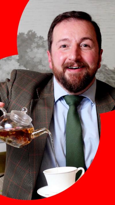 Man with brown hair and beard, in brown jacket and green tie, pours tea