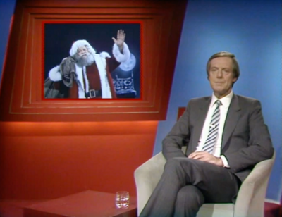 Barry Norman sits on chair with Santa on TV screen behind him