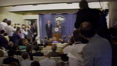 The view from the back of the White House press conference room.  Reports and cameras are in the foreground.  In the background is the President and his Press Secretary