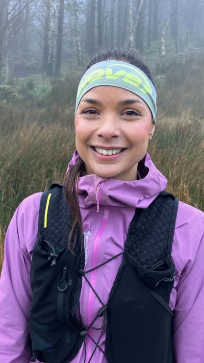 Sarah Perry. She is smiling and wearing a black running bag over a purple jacket. Her brown hair is tied back and she is wearing a headband. Behind her are trees and grassy bushes.