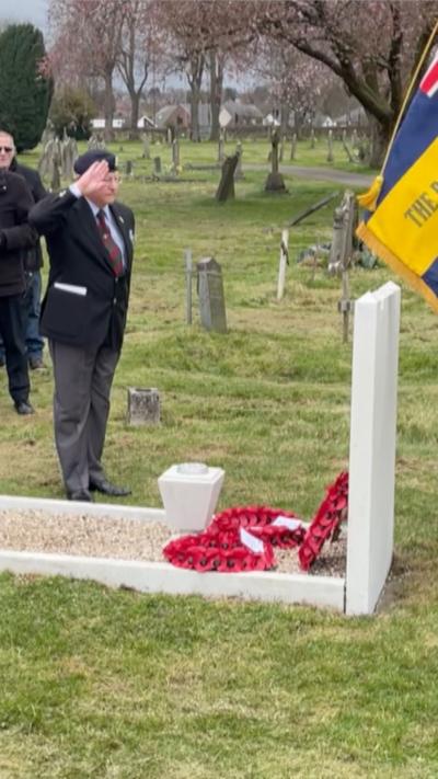 A person in formal clothing stands in a cemetery, raising a hand in a salute toward a grave adorned with red poppy wreaths, with a ceremonial flag visible in the foreground.
