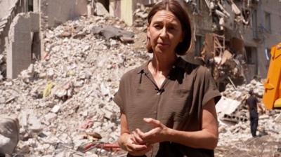 Sarah Rainsford wearing a bown short sleevd shirt standing in front of rubble after attacks in Kyiv