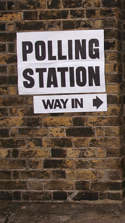 "Polling station" and "way in" is printed in black on white paper and has been stuck to brick wall.
