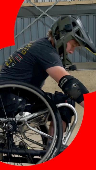 A side view of a teenager in a wheelchair, performing a stunt at a skate park, wearing a black helmet.