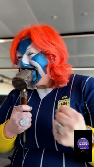 Michelle McManus - woman speaking into an old microphone with a saltire flag painted on her face, wearing a Scotland football shirt and shaking her fist with excitement
