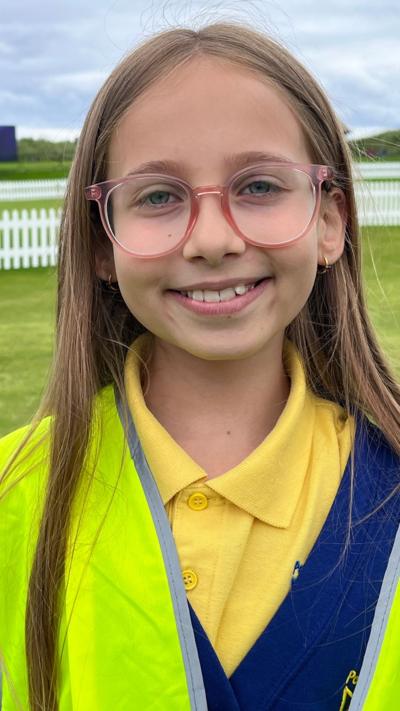 A young school girl with fair hair and glasses, wearing a yellow top and a high visibility vest.  She is standing on a golf course.
