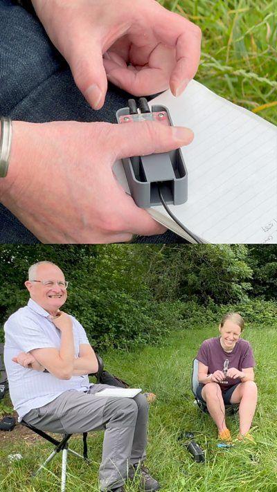 A split image, the top image shows a man's hands with a notepad a grey box with a wire coming out of it. The bottom image shows a man and a woman say in two fold-up deckchairs, sat in a field with hedges in the background. The man is wearing a pale blue shirt, grey trousers and glasses, the woman is wearing a purple t-shirt and black shorts. Both are smiling 