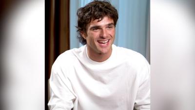 A man, Jacob Elordi, has dark brown hair and wears a white top. He is smiling