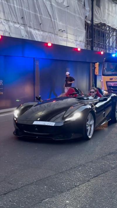 Black convertible sports car with red seats drives on city street near construction site and truck with flashing lights.