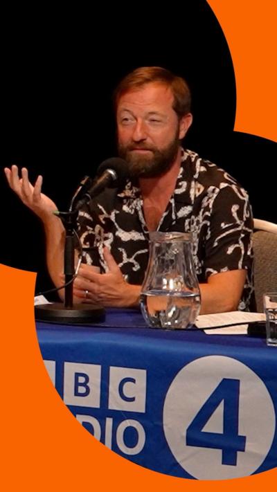 Irish comedian Andrew Maxwell on stage wearing a patterned shirt with his arm outstretched