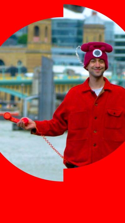 Joe Bloom is wearing a red phone-style hat, a red jacket and is carrying a red phone hook and cord, stood in front of a bridge.