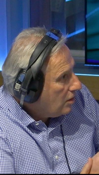 A man with grey hair, facing the right hand side of the picture and wearing black headphones and a blue and white checked shirt