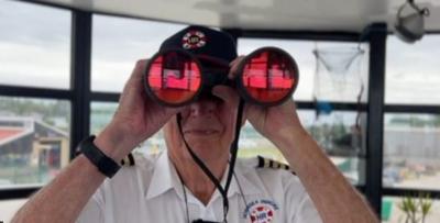 A man is holding a large pair of binoculars up to his eyes, he is wearing the uniform cap and shirt of Hornsea Inshore Rescue. 