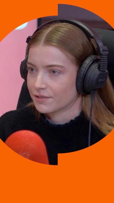 A woman with long brown hair wearing a black top sits in a radio studio in front of a microphone