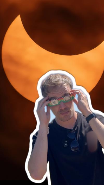Man wearing a black tshirt and paper solar viewing glasses with a photo of a solar eclipse behind
