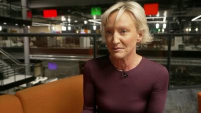 Ruth Dodsworth in the BBC studios in Cardiff. She is sitting on an orange sofa. She has cropped blonde hair and is wearing a fitted mauve top,