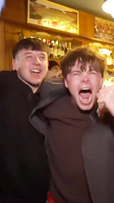 Two young men jump for joy as two older men are seen hugging next to them in a pub