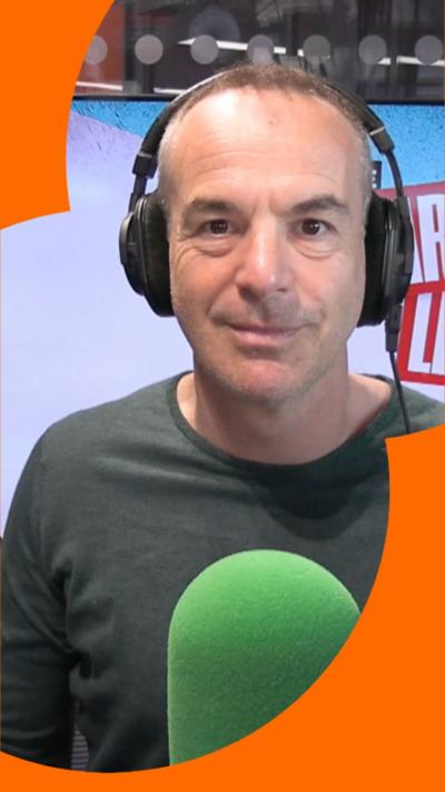 A man with short hair stands in front of a microphone, he smiles at the camera
