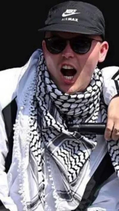 A kneecap member wears a cap and keffiyeh. 