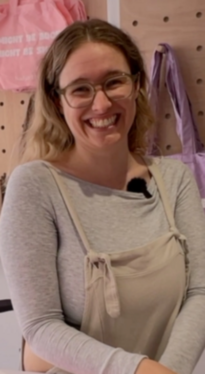 Woman with brown hair and glasses wearing a grey top and grey dungarees smiles at the camera