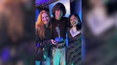 A woman with ginger hair, a black leather jacket and blue jeans holds a BBC Introducing Artist of the Year trophy. She is standing beside a person with black curly hair, wearing a gold necklace, a dark jumper and light grey jeans. They are standing next to another woman who has black hair and is wearing large round earrings, a white top, a tartan dress and a silver chunky necklace. They are standing in a BBC Scotland radio studio.