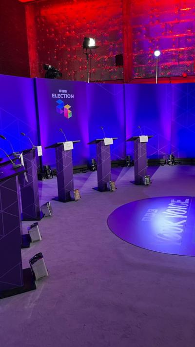 An empty studio with BBC Election 2026 branding lit up in purple with six empty lecterns. The top of the screen is lit up in red