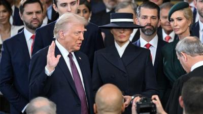 Donald Trump stands with his right hand raised and Melania at his side