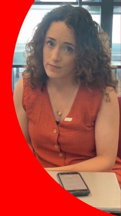A woman with long, curly hair looks at the camera. She wears an orange sleeveless top.