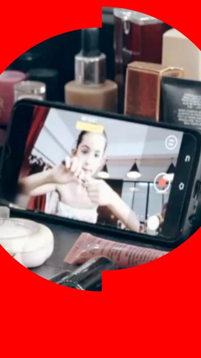 Young girl is seen sat in front of a phone screen that's recording her. She's pulling open a tube of makeup with her hands, and the phone is surrounded by various makeup products