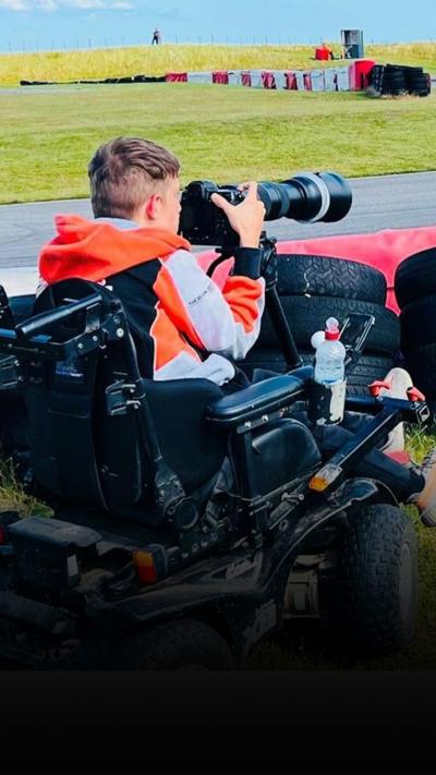Jake Smith - man in electric wheelchair, holding a long lens camera at a racetrack