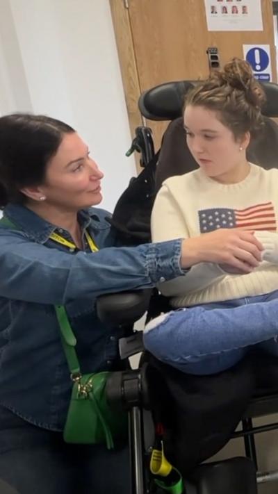 A woman with dark hair and a blue denim shirt squatting down by a girl in an electric wheelchair.