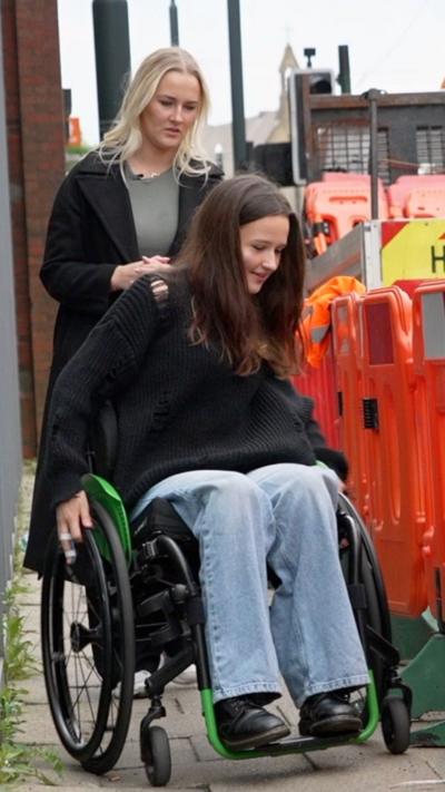 A woman using a wheelchair along a pavement beside orange roadwork barriers with another individual.