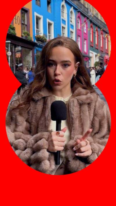 A woman in a brown woolly coat stands in an Edinburgh street speaking into a handheld mic. There is a row of multicoloured houses behind her