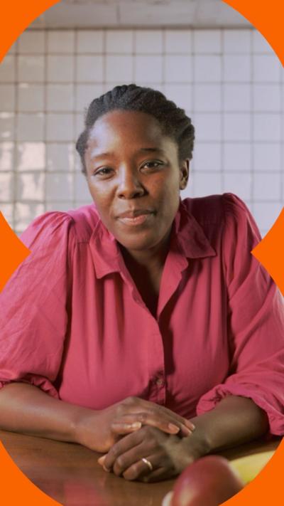 A woman with tied-back black hair, sits in a kitchen wearing a pink shirt