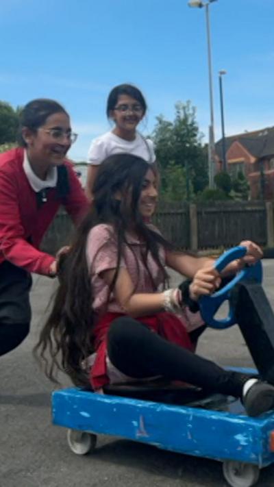 Two girls push another girl around a playground on a blue and black wooden go-kart