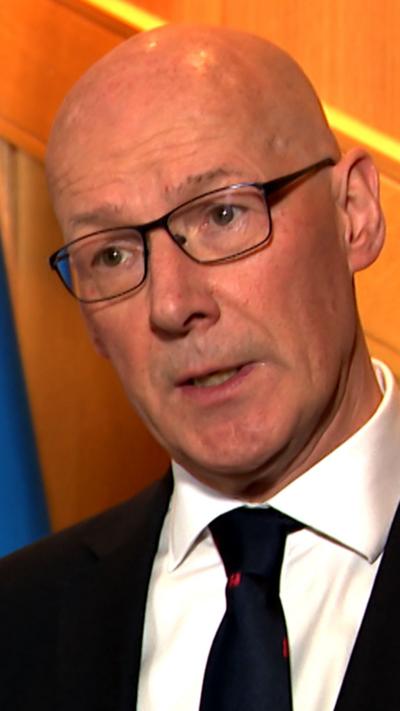 John Swinney standing beside a Saltire, he is bald, has glasses and a dark suit and tie.