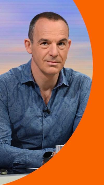 A man, Martin Lewis, looks pensive sat in a studio. He has short dark hair and wears a blue shirt 