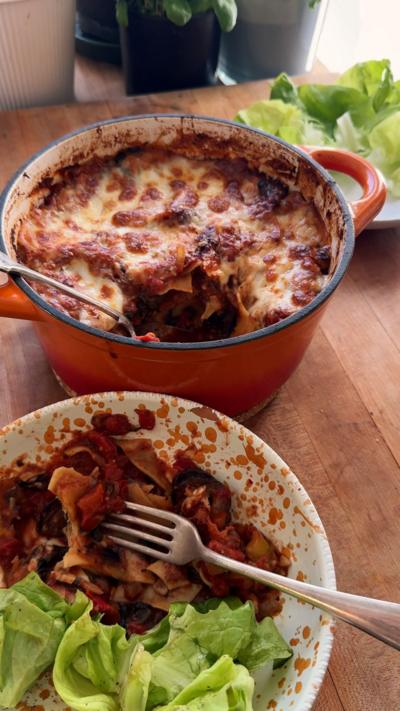 A large pot filled with a lasagne, with a bowl of one portion served with lettuce on a wooden worktop.