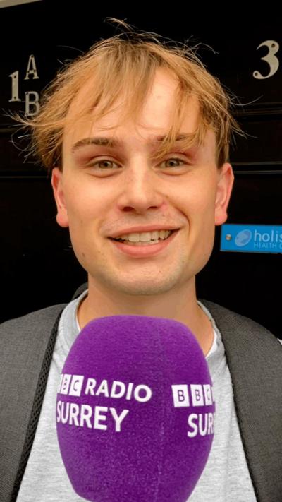 A young man with long, brown hair standing in front of a purple microphone.