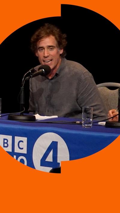 Stephen Mangan speaks into a microphone. He has brown hair and wears a grey shirt